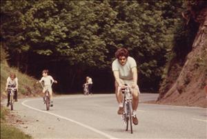 An adult and two children ride bikes up a steep curving road. Behind them, a person walks their bike up the hill. 